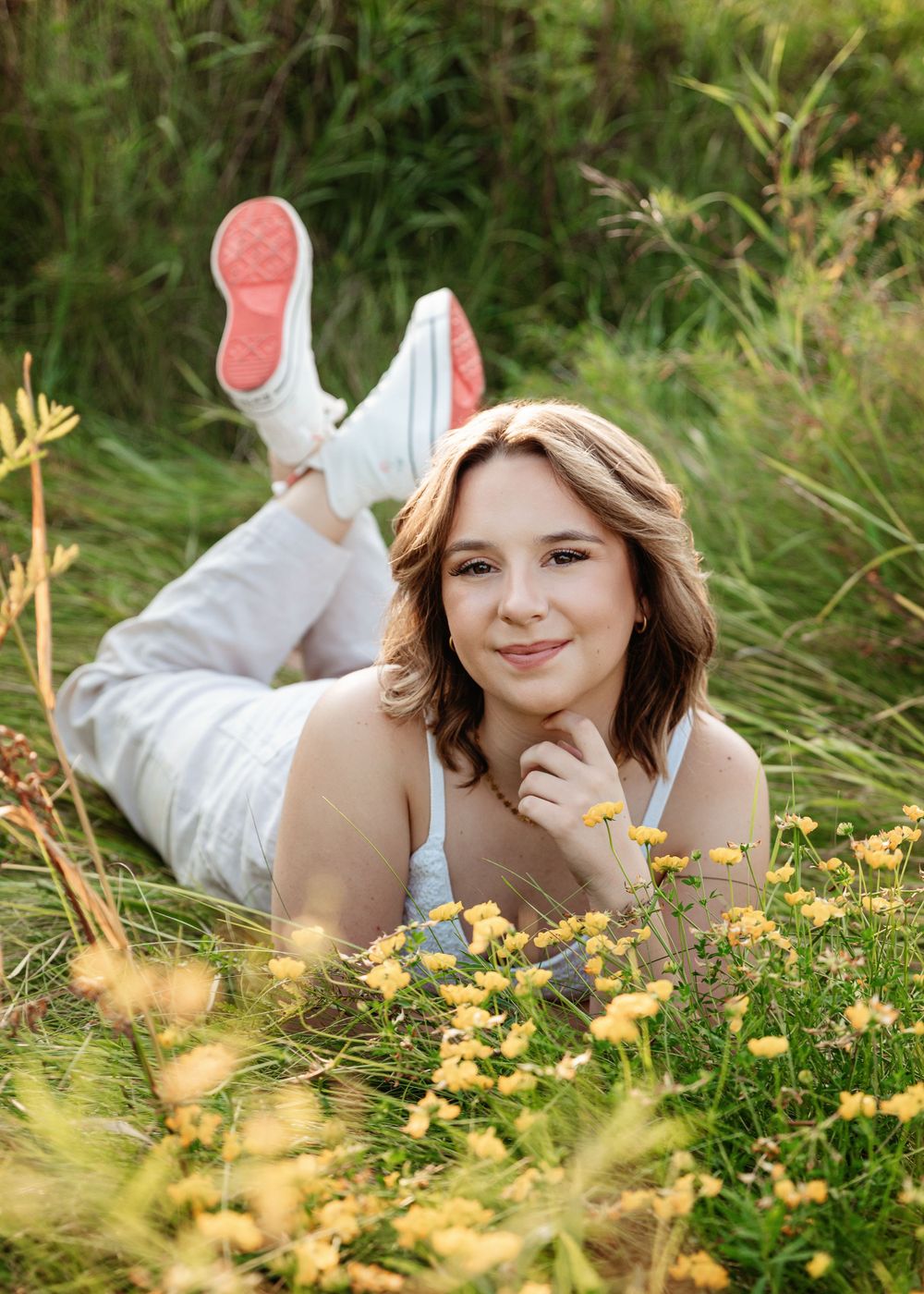 A figure lies in a lush meadow with yellow wildflowers, wearing white clothing and pink sneakers on a sunny day.
