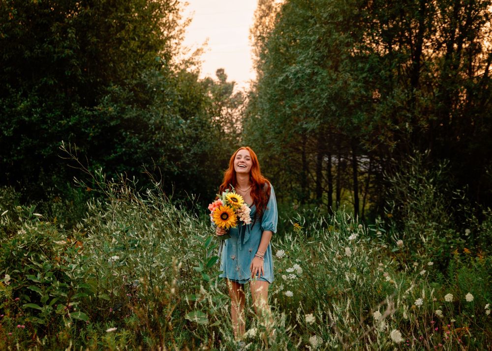 A figure in a blue dress holds bright sunflowers while standing in a wildflower meadow at sunset.