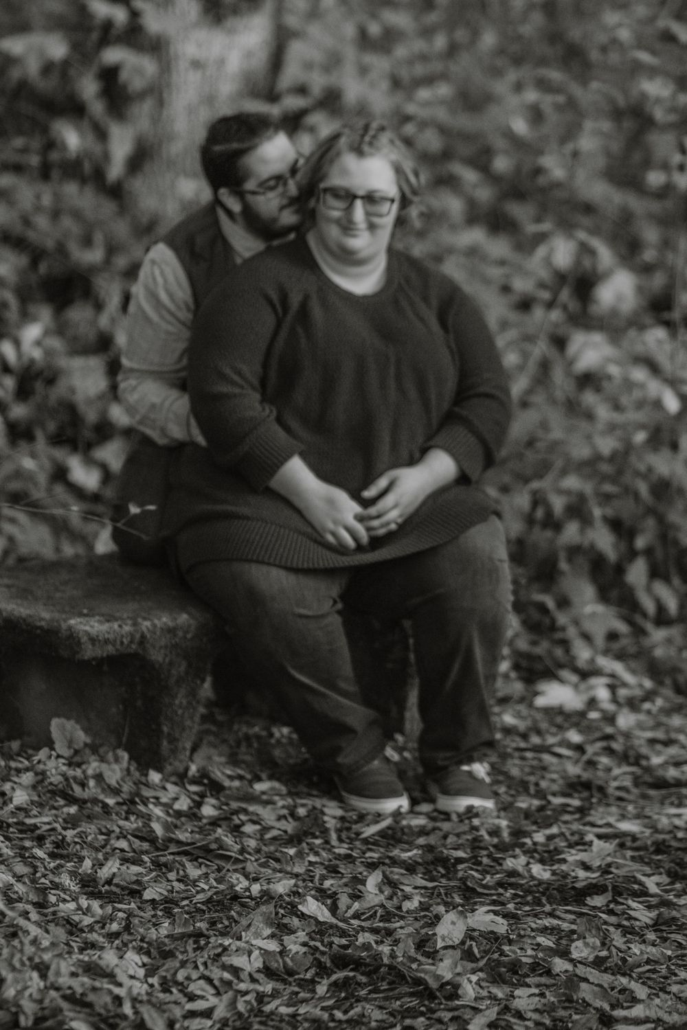 Black and white engagement photo of couple sitting together on stone bench in autumn woods.