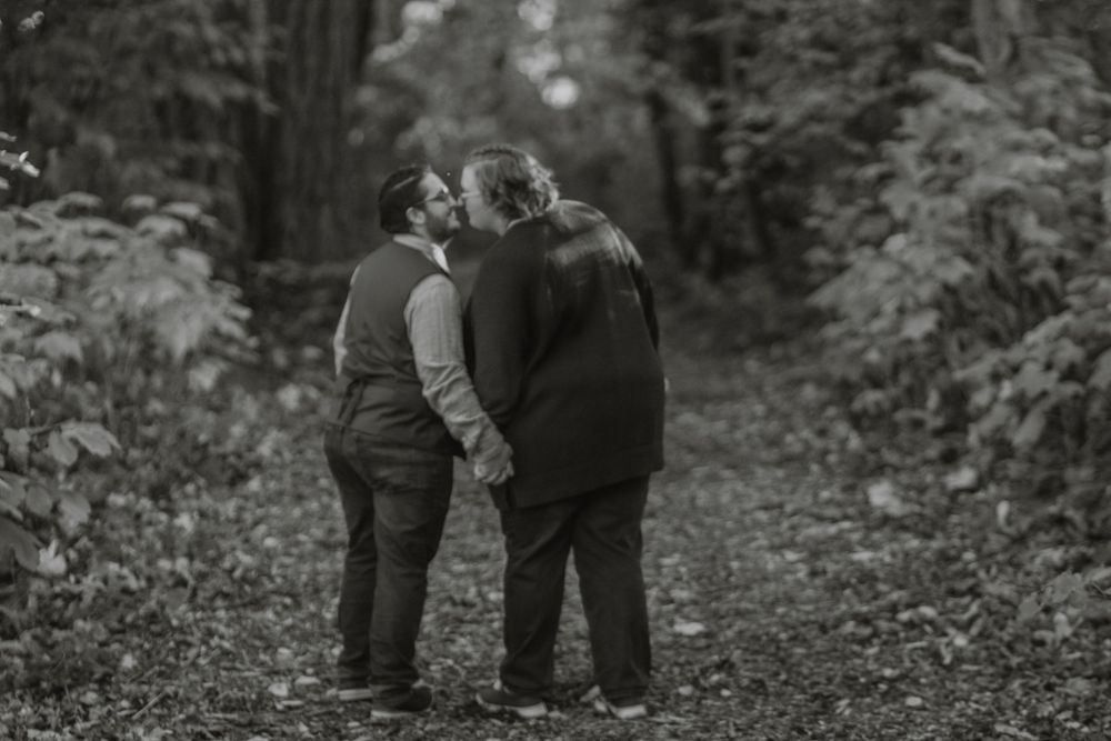 Black and white photo of couple kissing on a leaf-covered forest path holding hands.