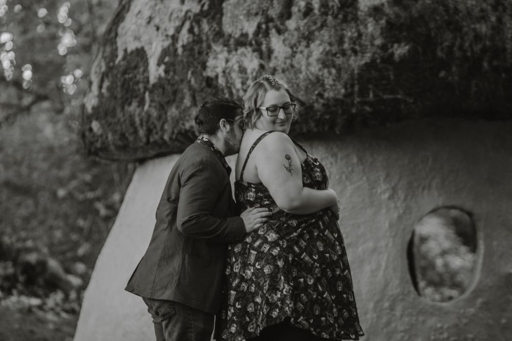 Black and white photo of a couple sharing a tender moment against a rustic stone wall outdoors.