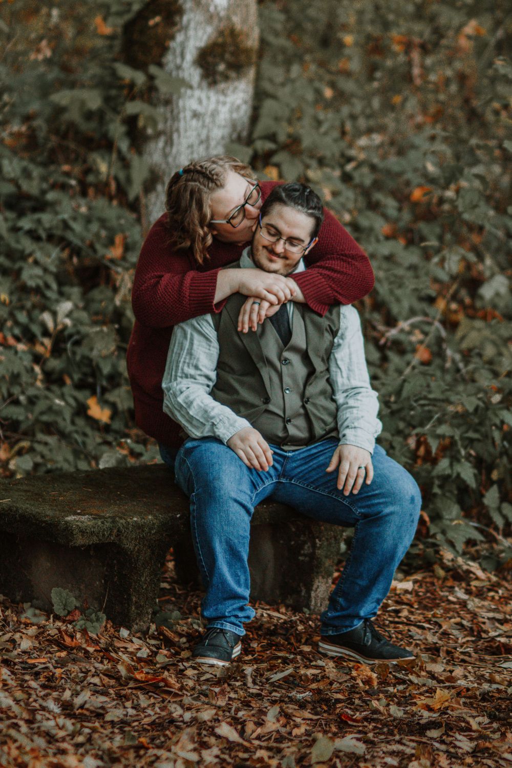 Couple sharing tender moment in autumn forest, woman kissing mans cheek as he sits on stone bench.