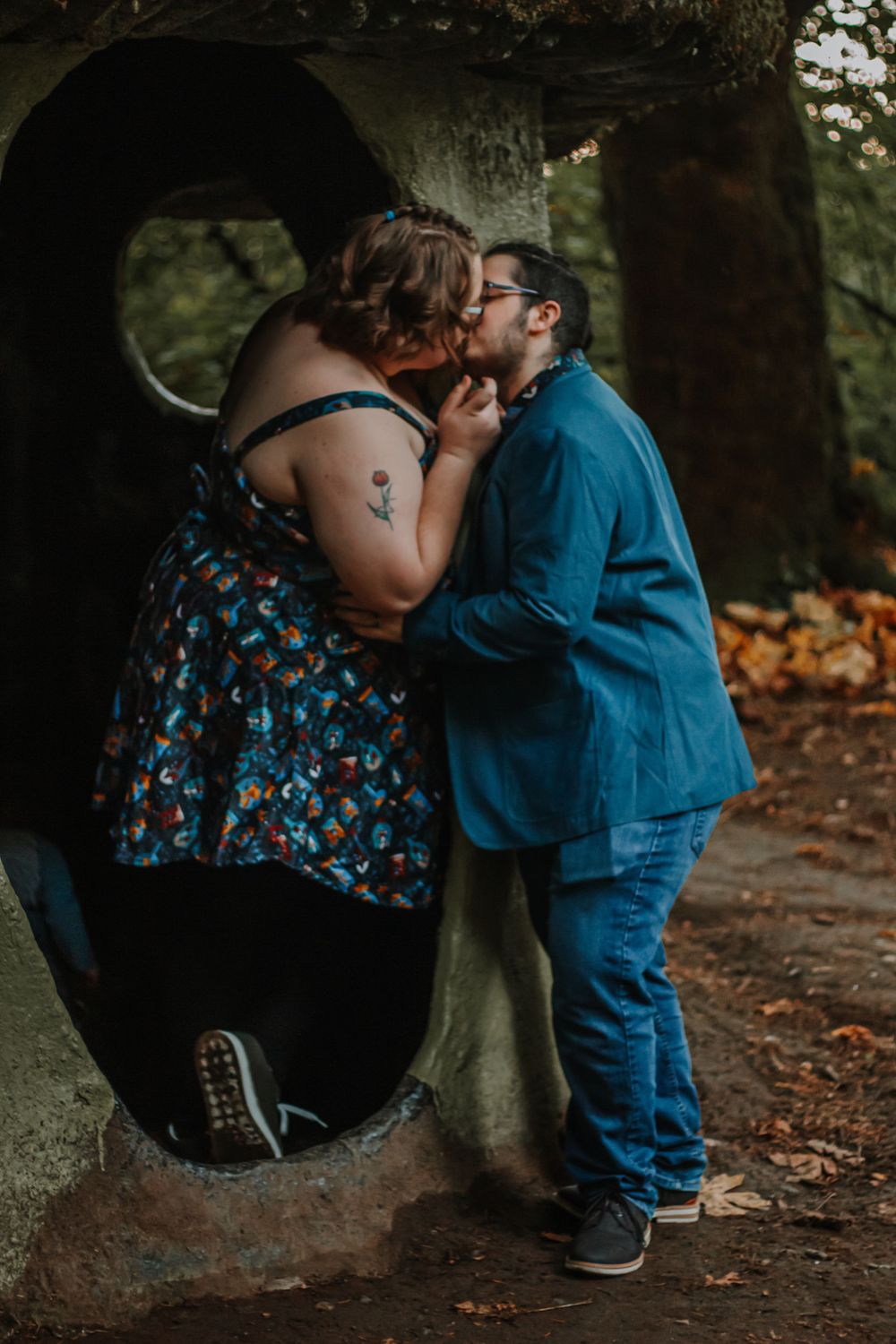 A couple shares a romantic kiss near a large stone sculpture in a wooded park.