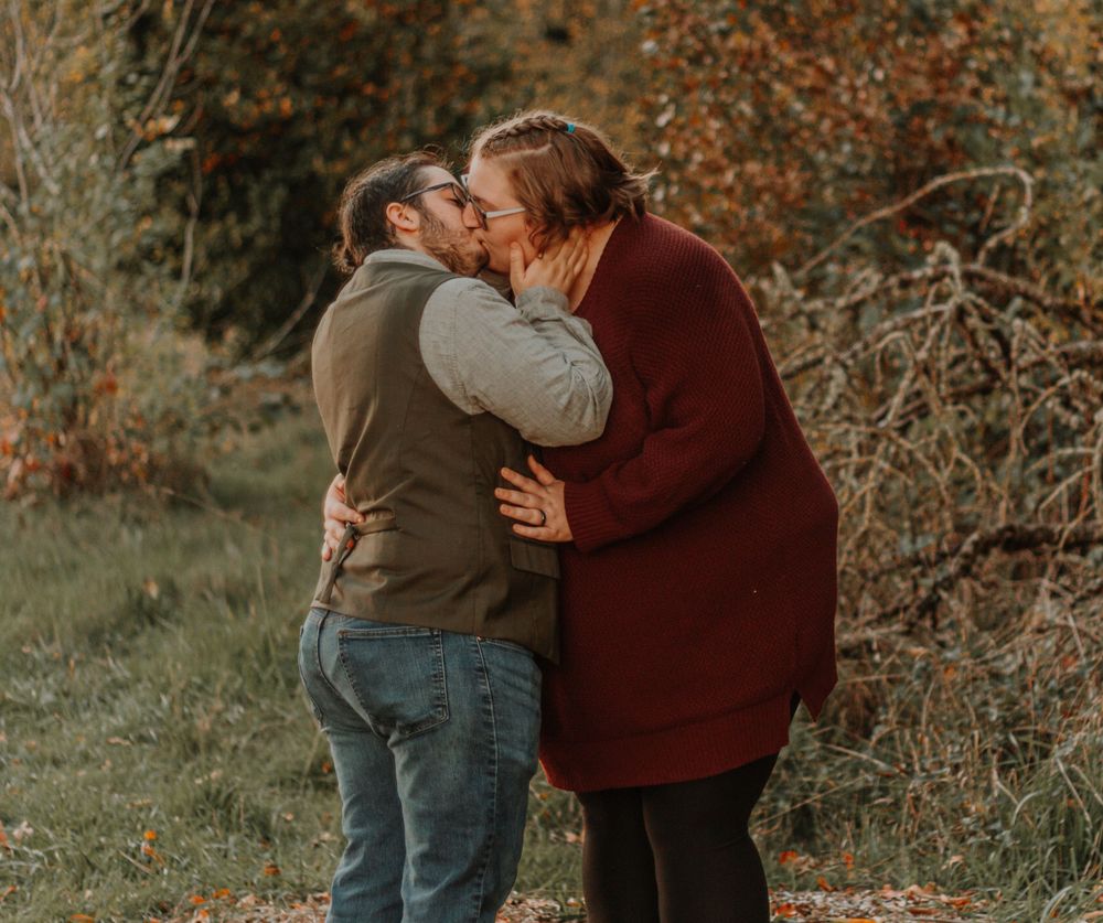 A couple shares a loving kiss outdoors surrounded by autumn trees and foliage.