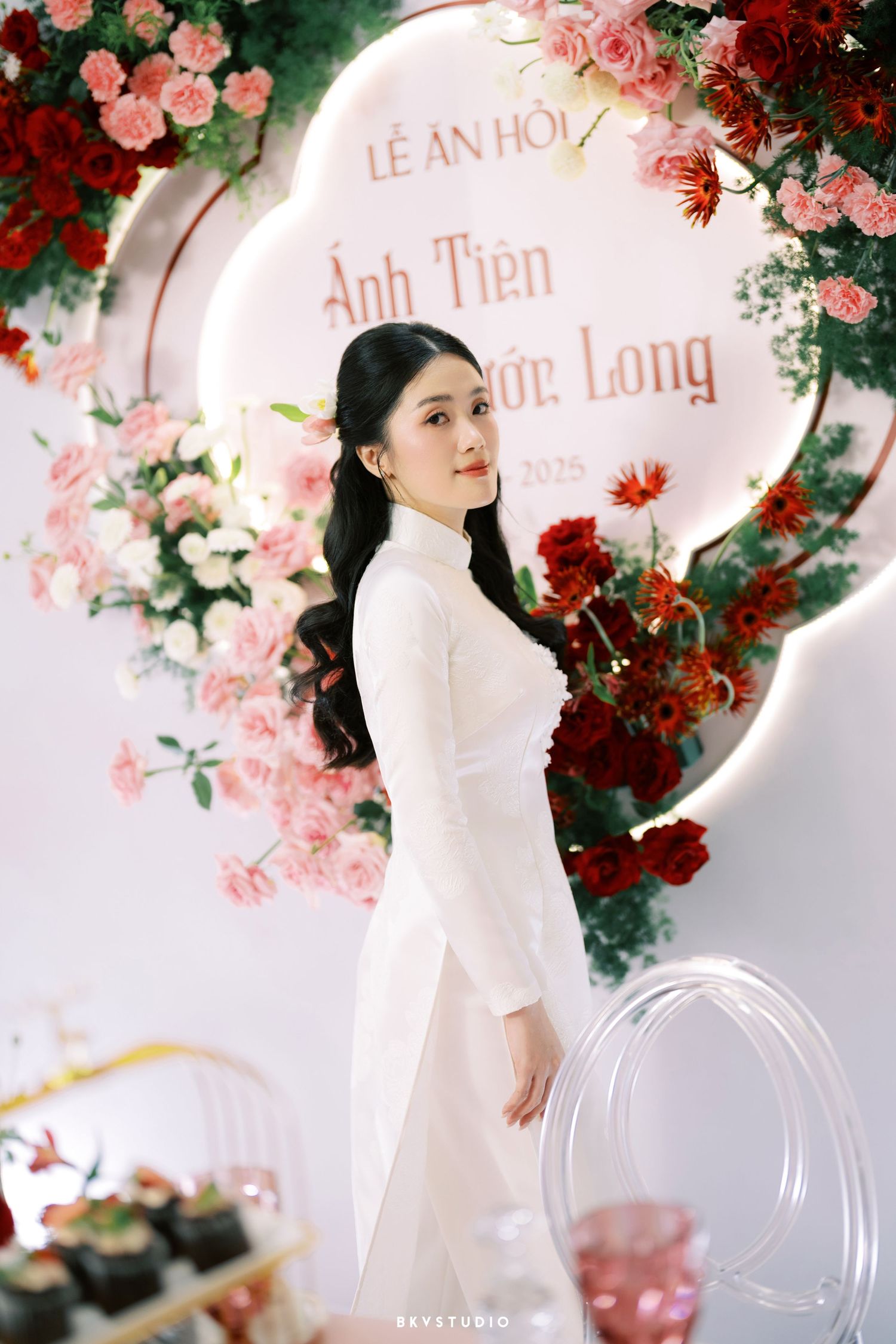 A traditional white ao dai dress in front of an elegant floral arch with pink and red roses for a wedding celebration.