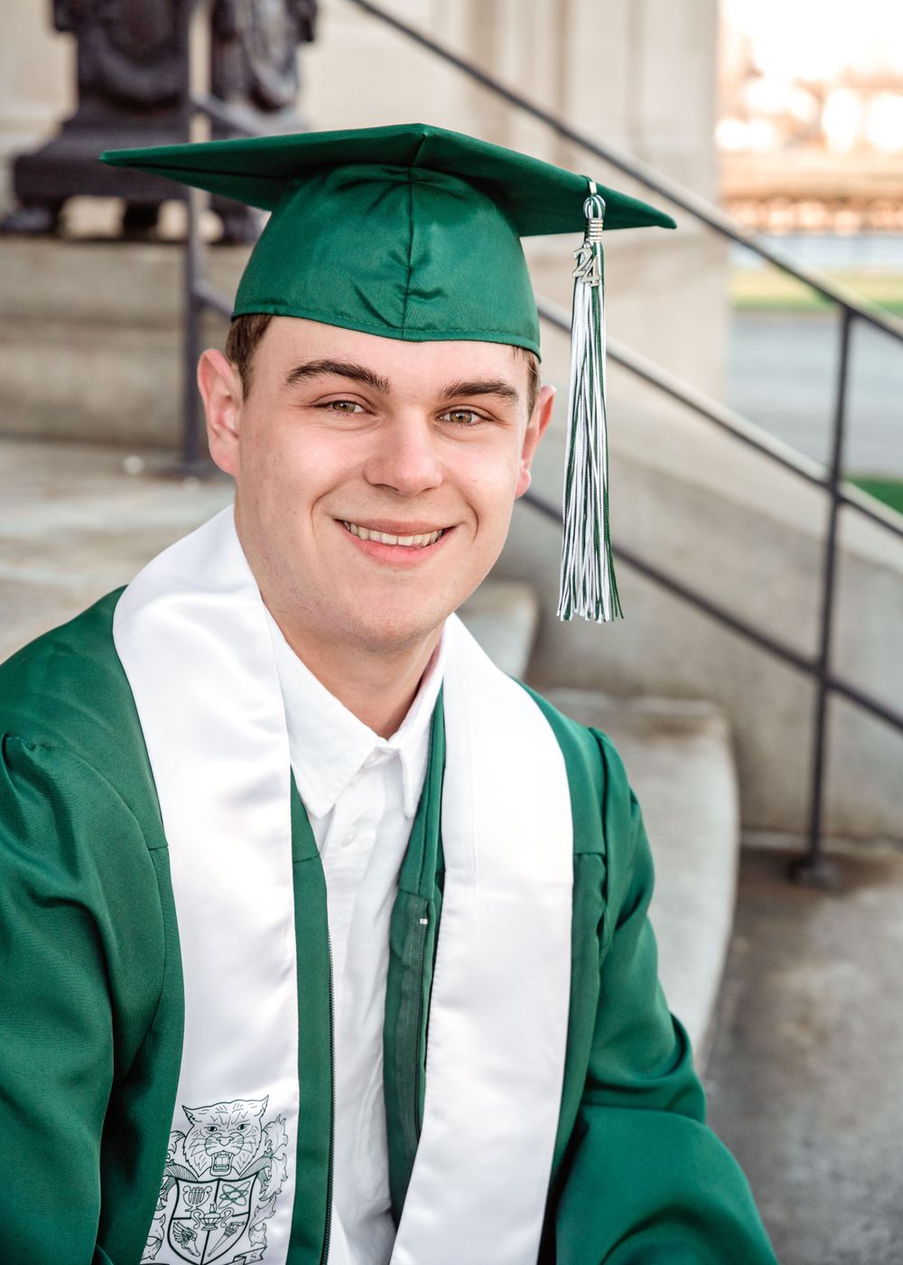 A graduate in green cap and gown with a silver tassel poses for a photo on concrete steps.