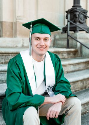 Graduate in emerald green cap and gown sits smiling on stone steps of a building during graduation.