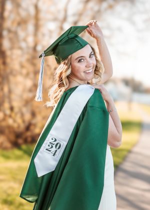 Graduate in emerald green cap and gown poses outdoors with a white sash during a sunny fall day.