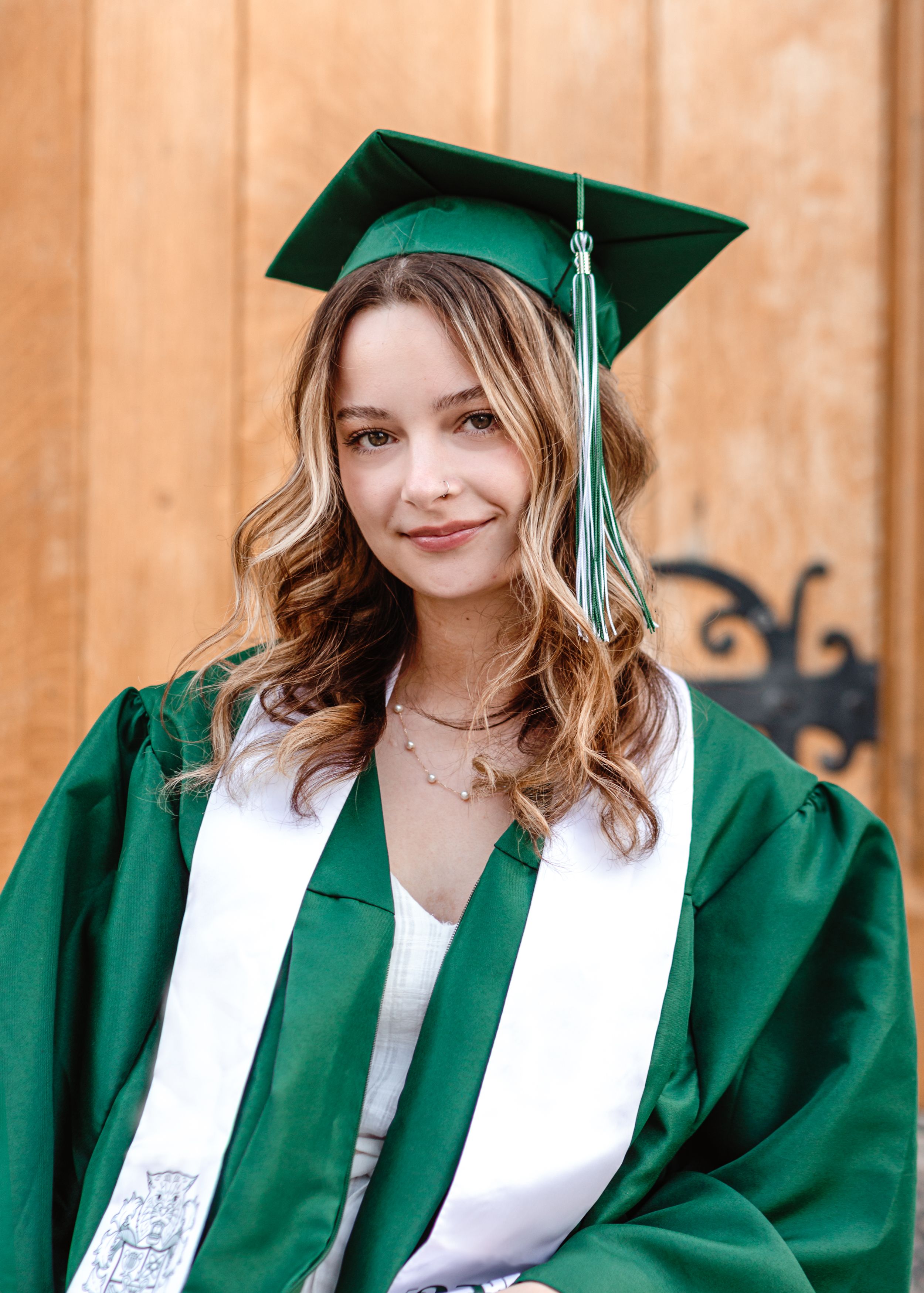 A graduate in green academic regalia and white stole with wavy hair poses against a wooden background.
