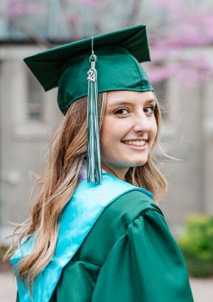 A graduate in an emerald green cap and gown with silver tassel smiles while posing against spring blossoms.