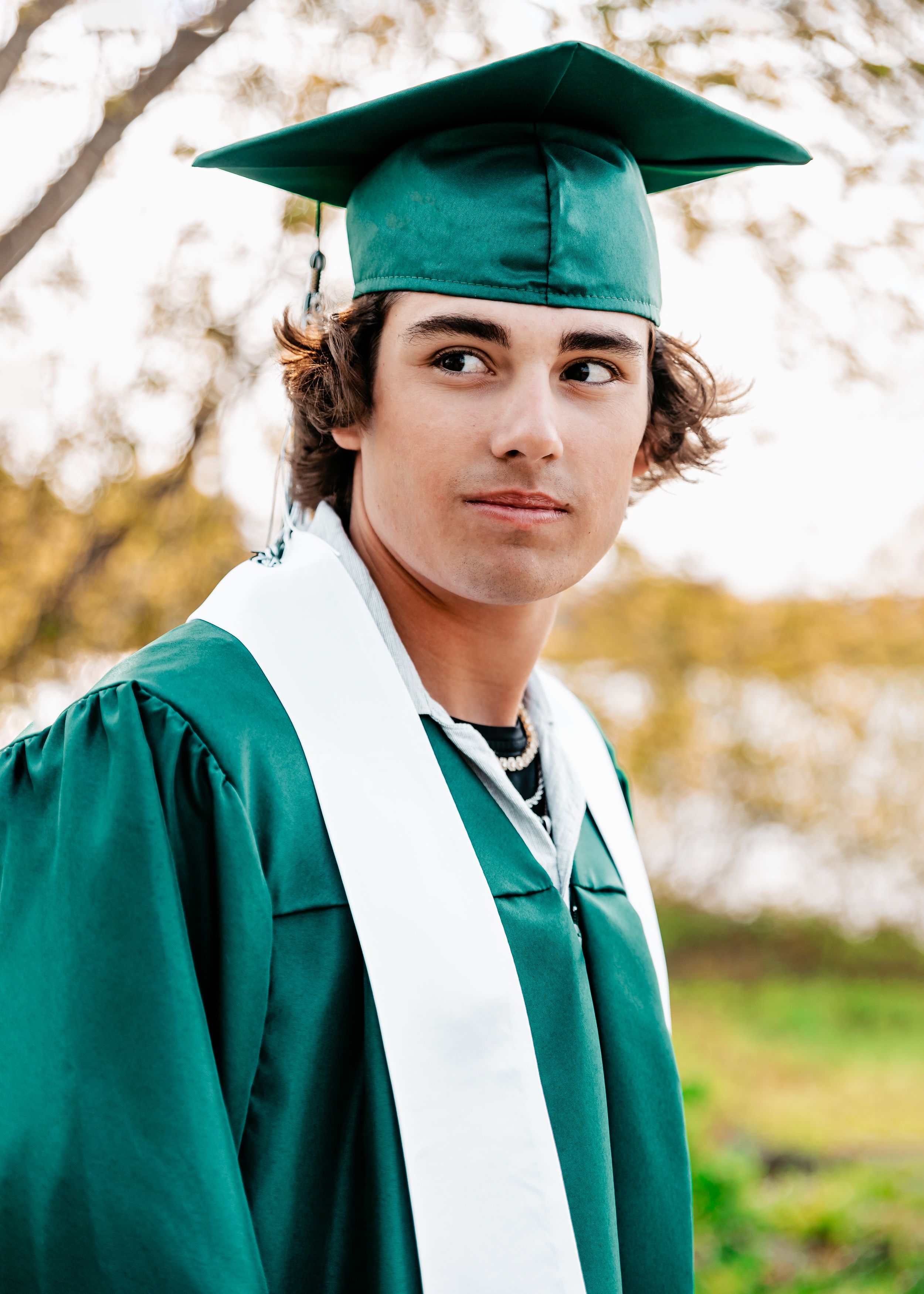 Graduate wearing emerald green cap and gown with white stole poses outdoors on sunny day for graduation photo.