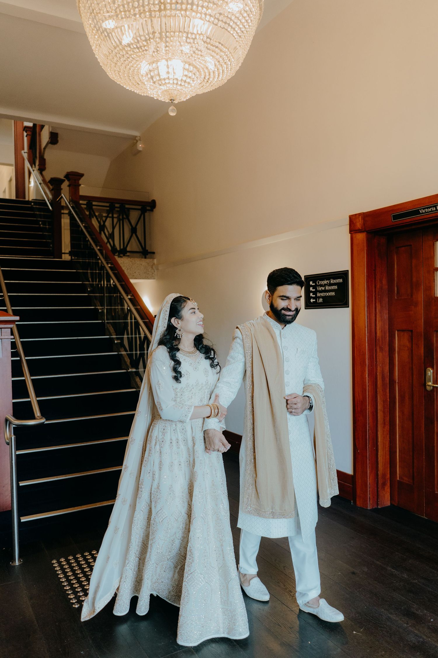 A couple in elegant ivory traditional outfits stand by a grand staircase under a crystal chandelier.