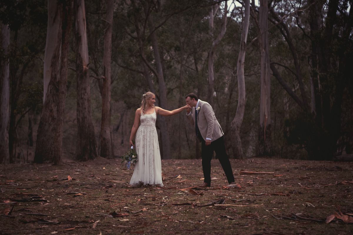A couple in formal attire is standing in a forest. The woman is wearing a white dress and holding a bouquet, while the man is in a suit, ben