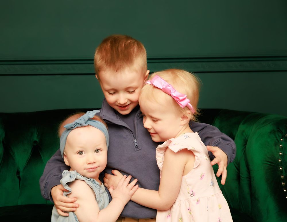 Three small children share a joyful moment together on a green velvet couch.