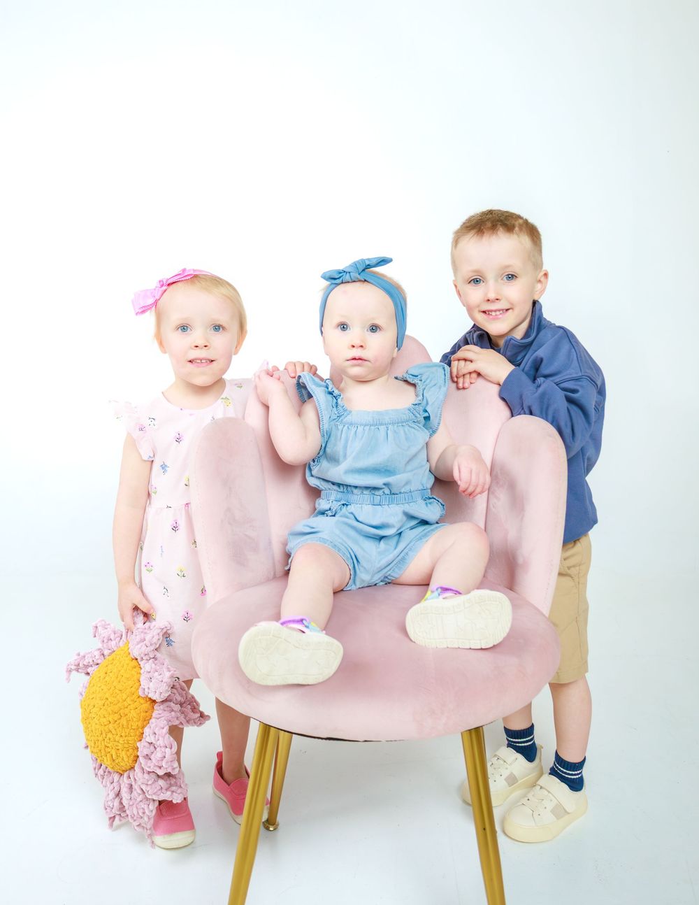 Three children in coordinated outfits pose together on a pink chair against white background.