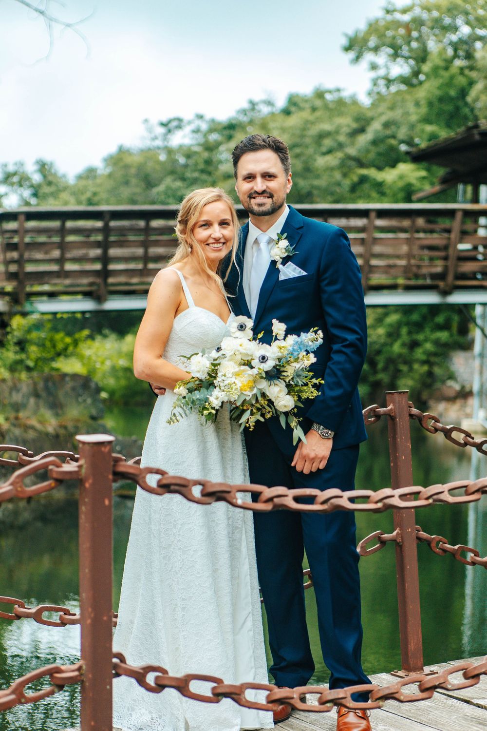 Newlyweds share joyful moment on wooden bridge with decorative chain railing near lake.