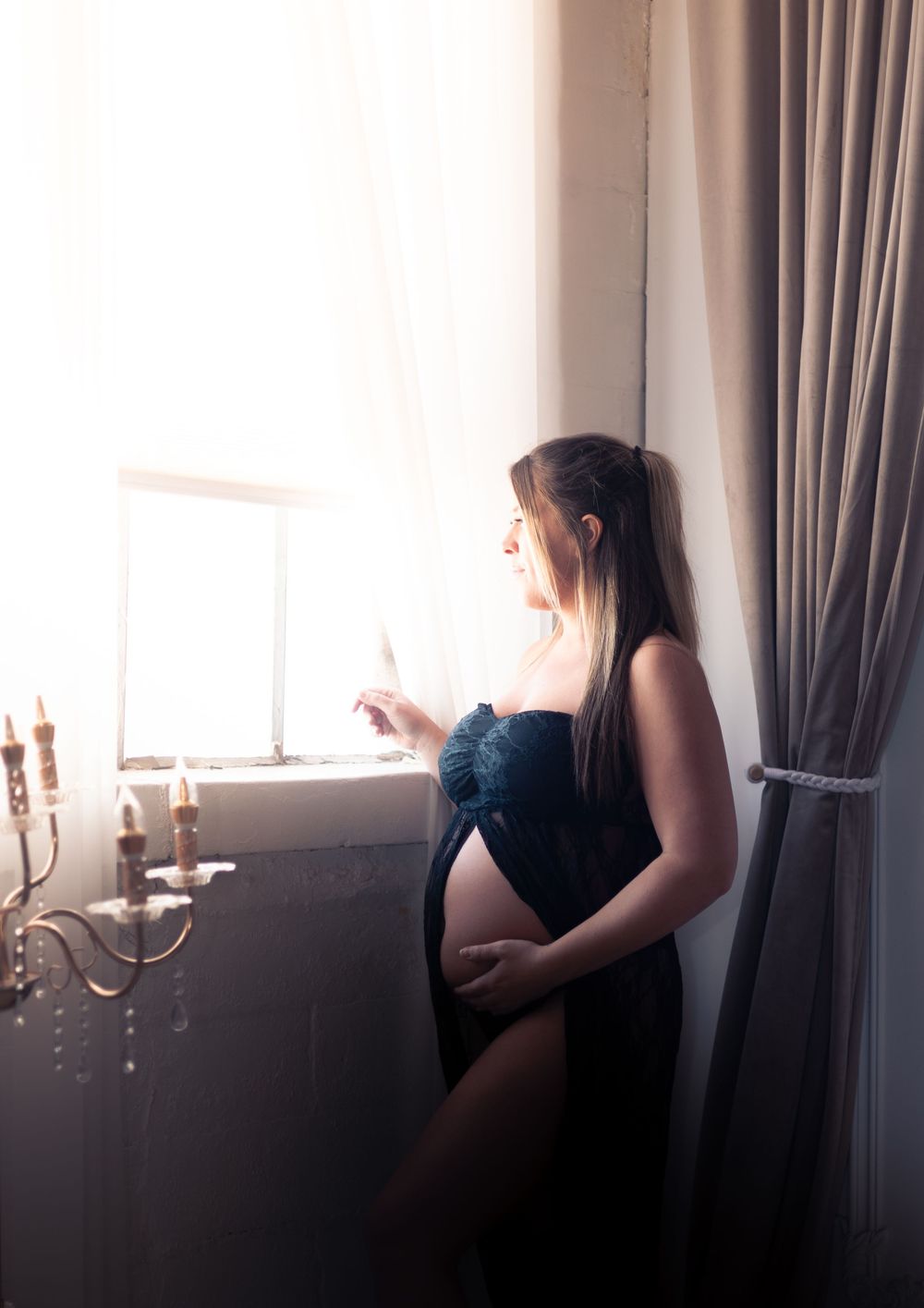 Silhouetted figure stands near window with elegant candelabra in moody indoor boudoir setting.
