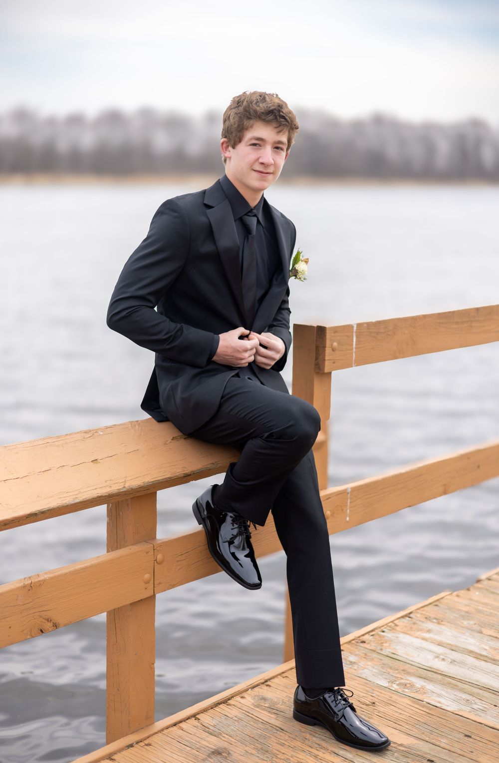 A formally dressed person poses on a wooden dock overlooking a lake.