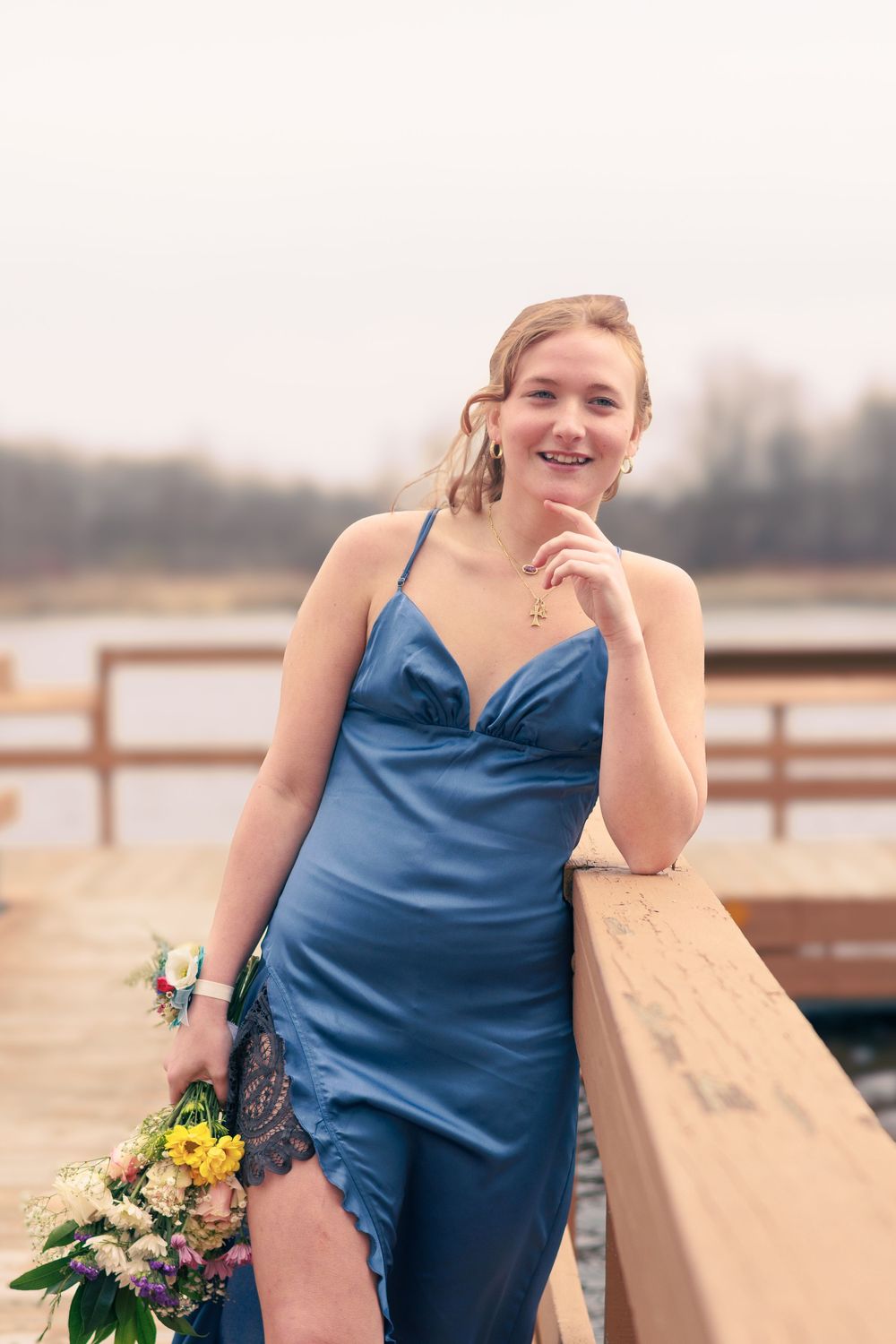A person in a blue silk dress poses with flowers on a wooden dock by the water.