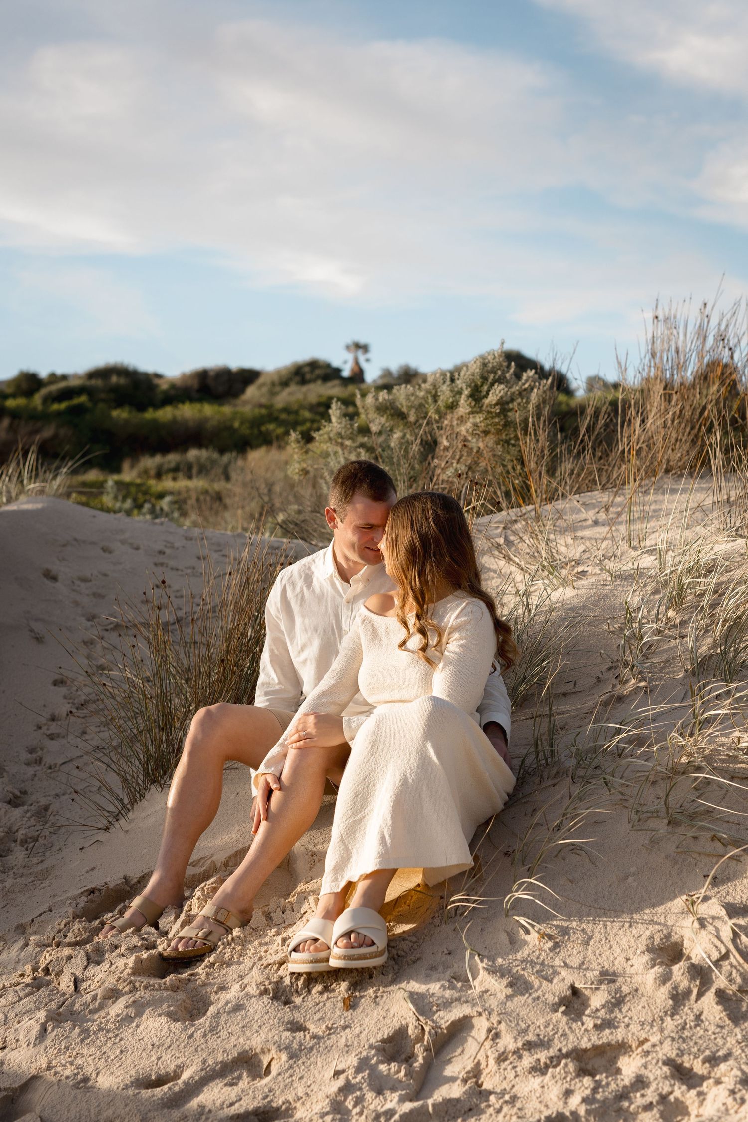 A romantic couple sits together on sandy beach dunes, dressed in white, sharing an intimate moment with coastal greenery behind them.
