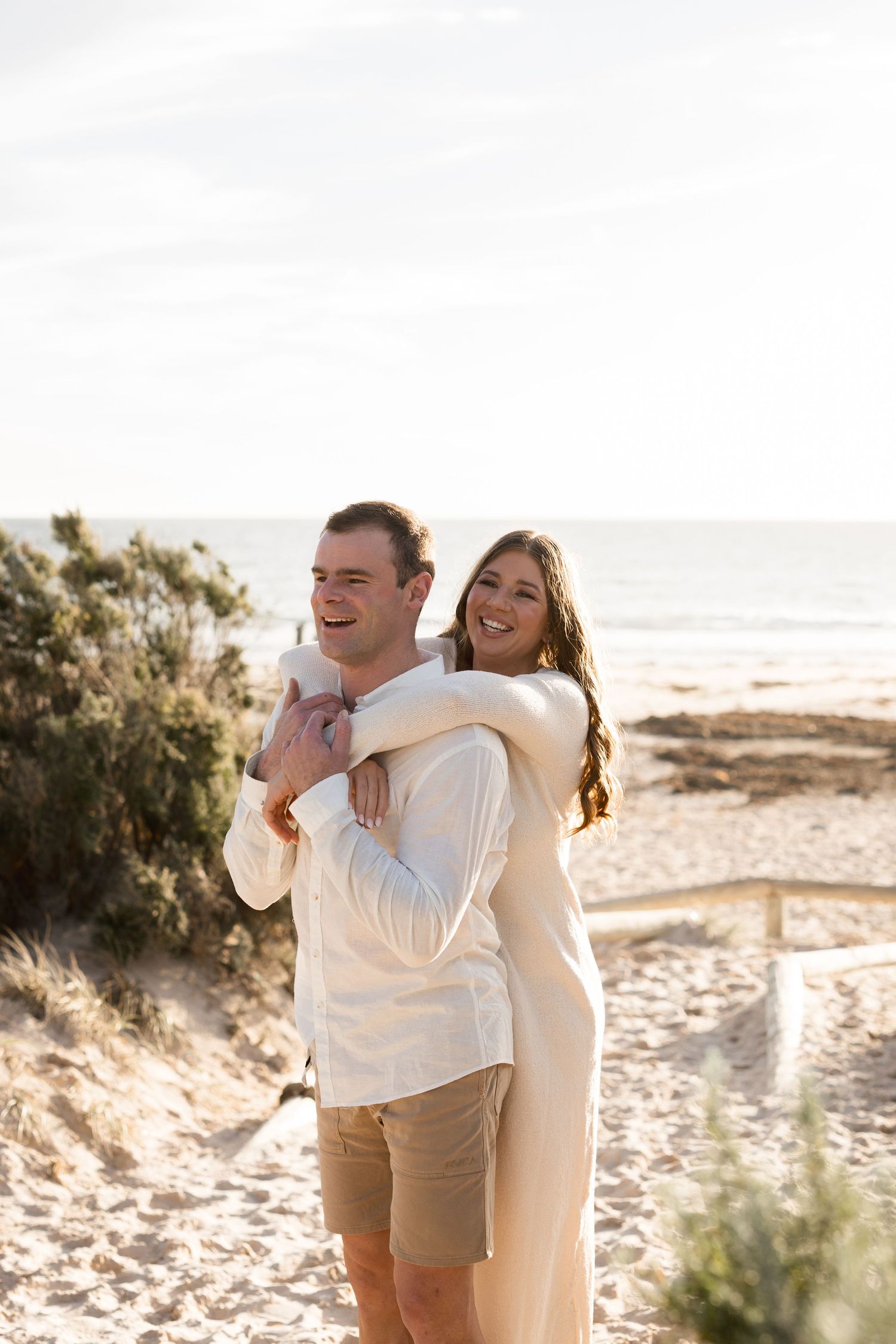 Happy couple embracing on a sandy beach path, smiling in white outfits with ocean backdrop.