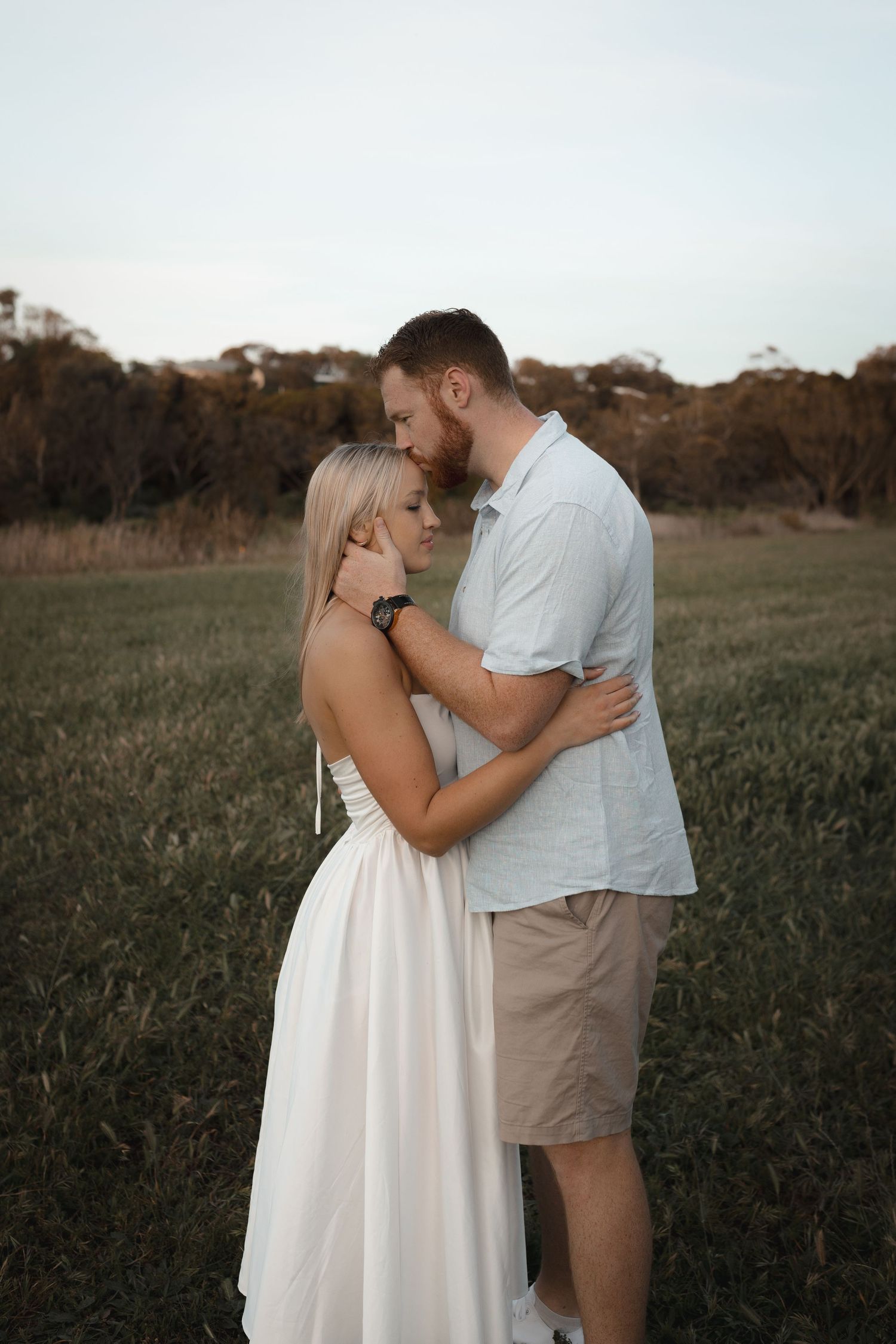 A couple embraces tenderly in a green field, woman in white dress, man in light shirt and shorts, trees in background.