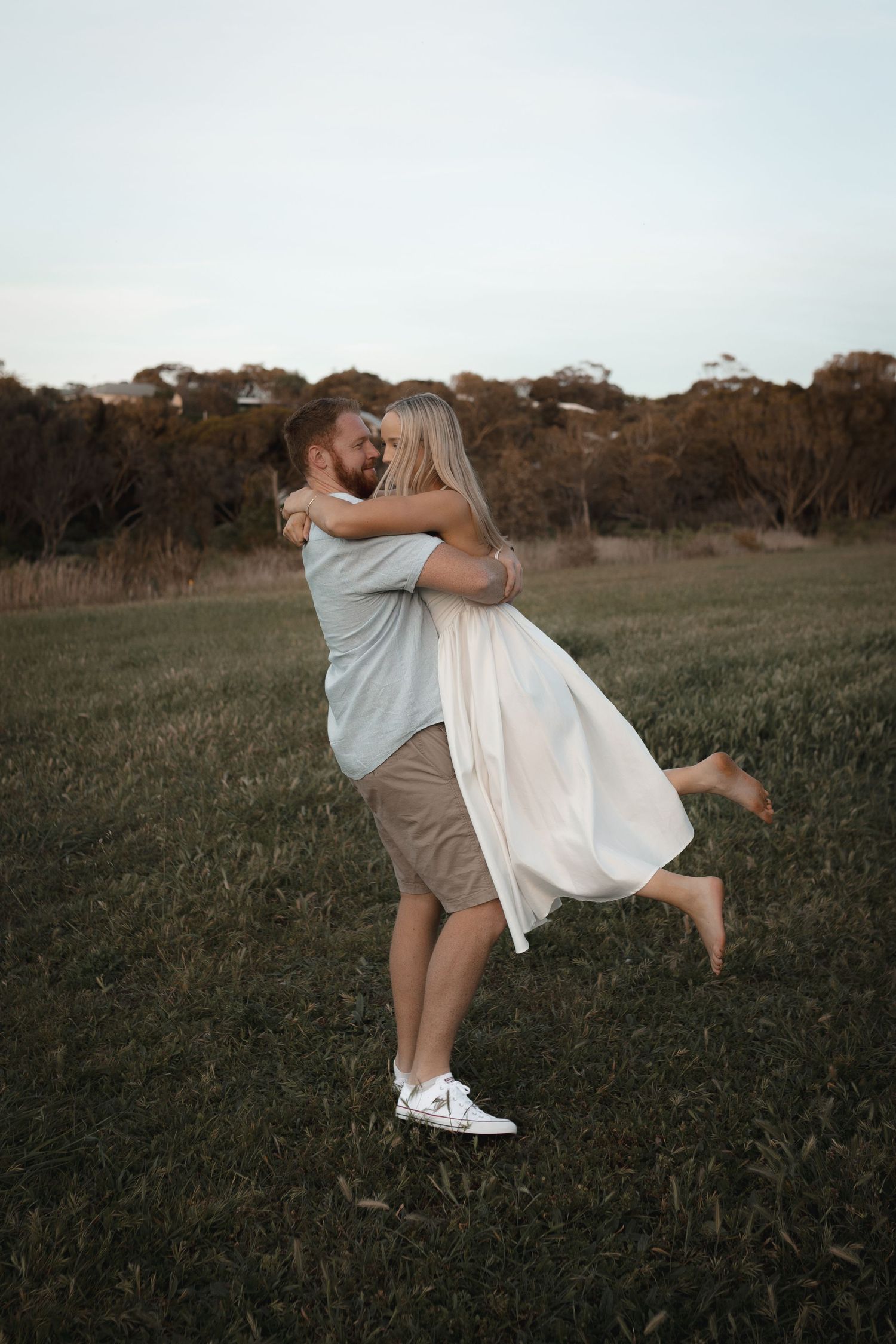 Couple embracing in a green field, man lifting woman in white dress, trees in background at dusk.
