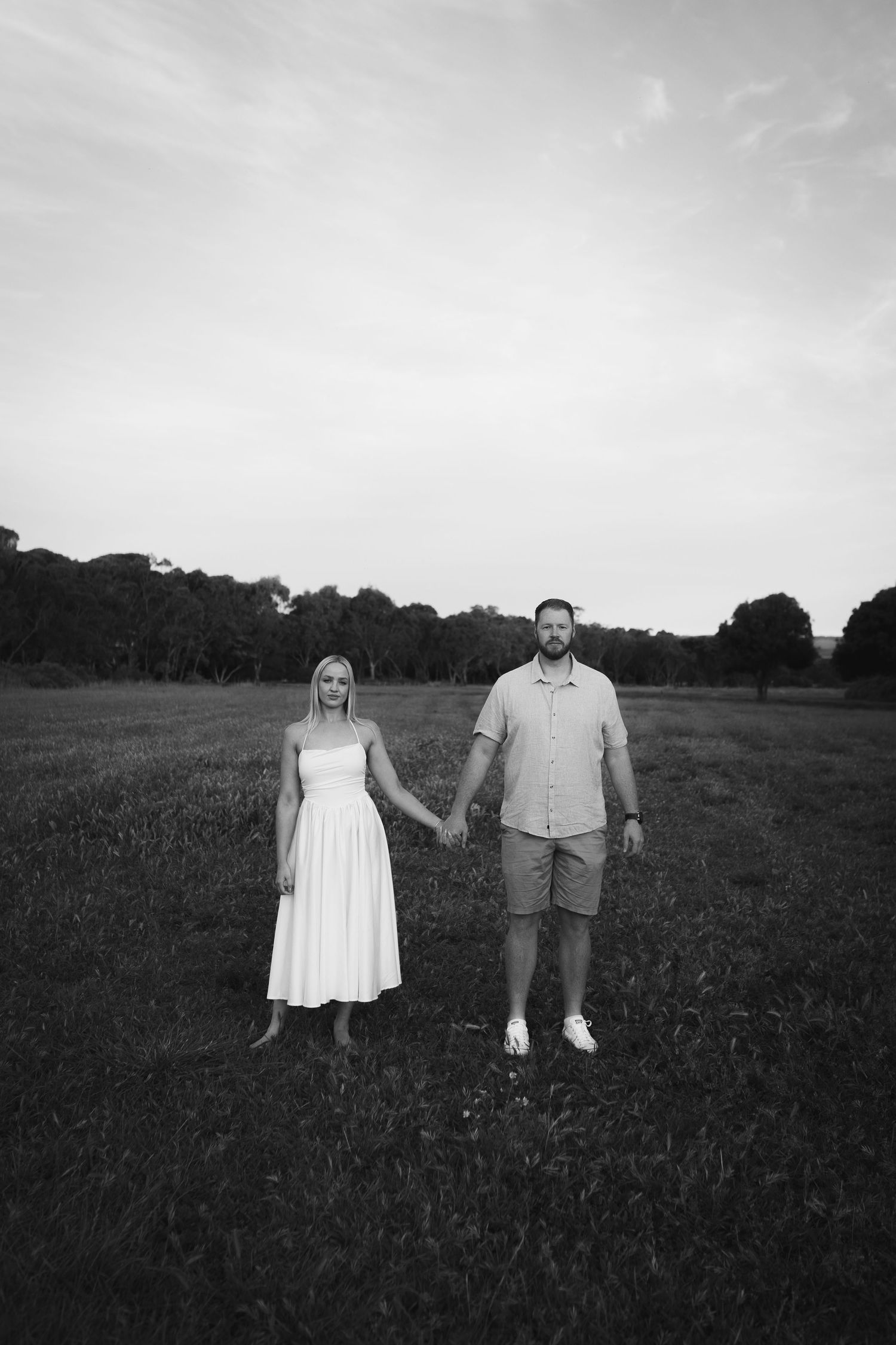 Black and white photo of couple holding hands in open field with trees and cloudy sky behind them.