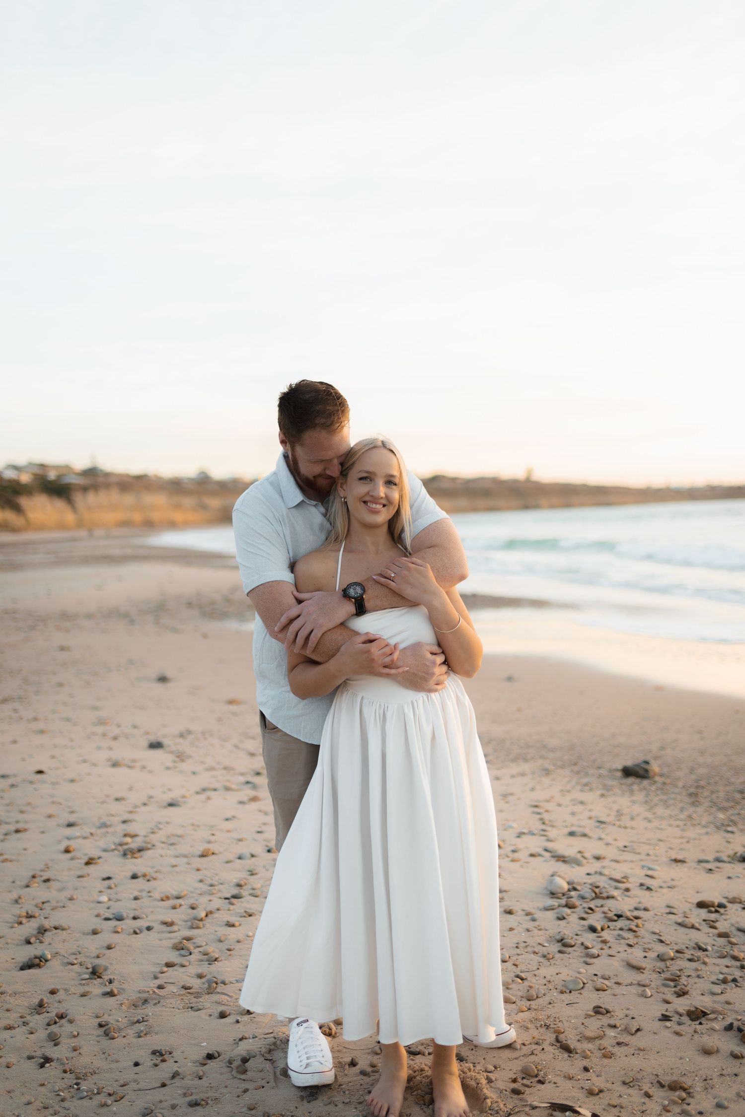Couple embracing on a sandy beach at sunset, woman in white dress smiling as man holds her from behind near the ocean.