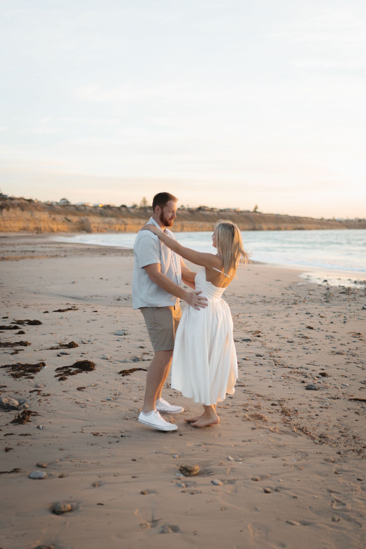 Couple dancing on a sandy beach at sunset, woman in white dress, man in white shirt and shorts, coastal cliffs in background.