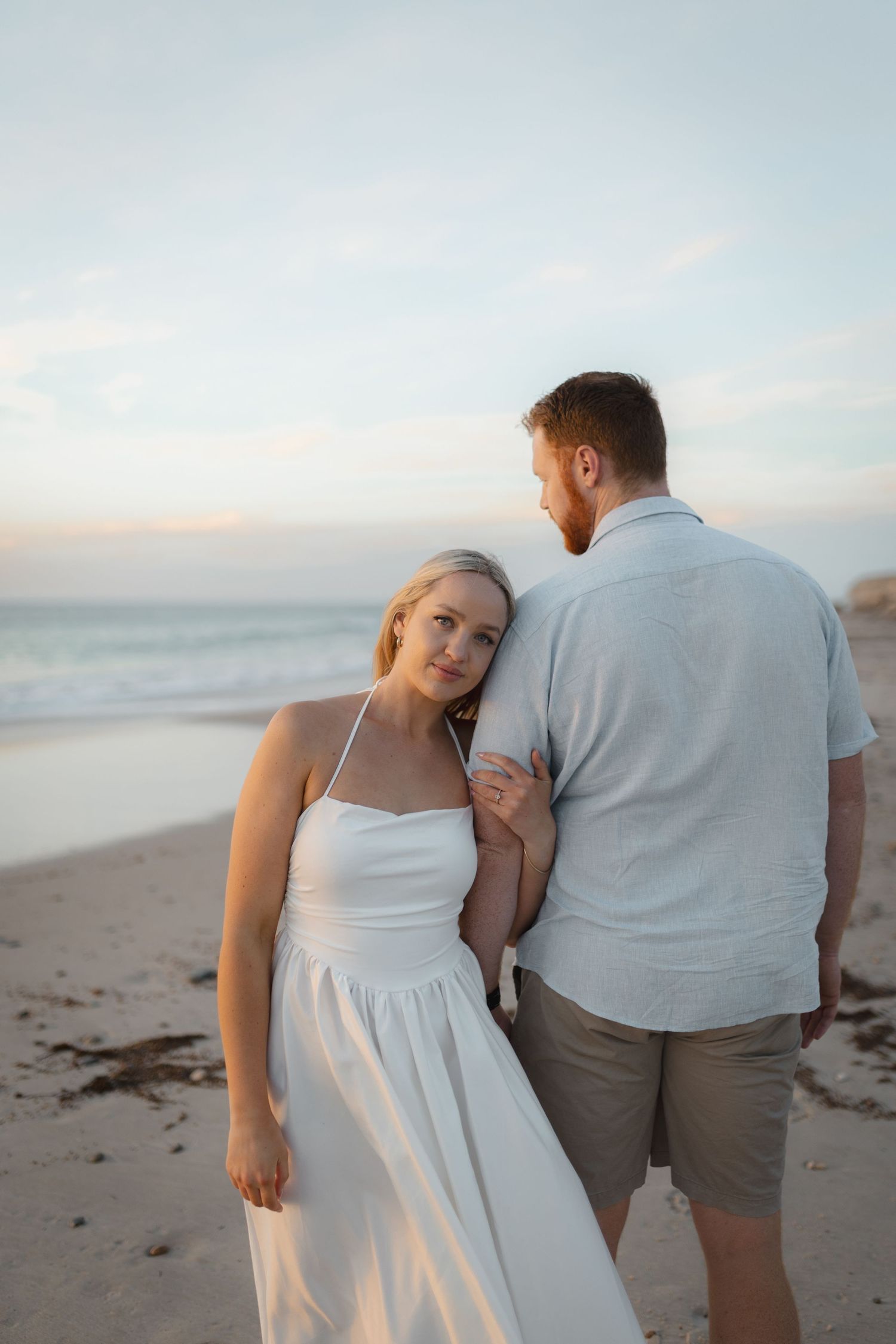 Woman in white sundress leans on mans shoulder at sunset beach, ocean waves in background.