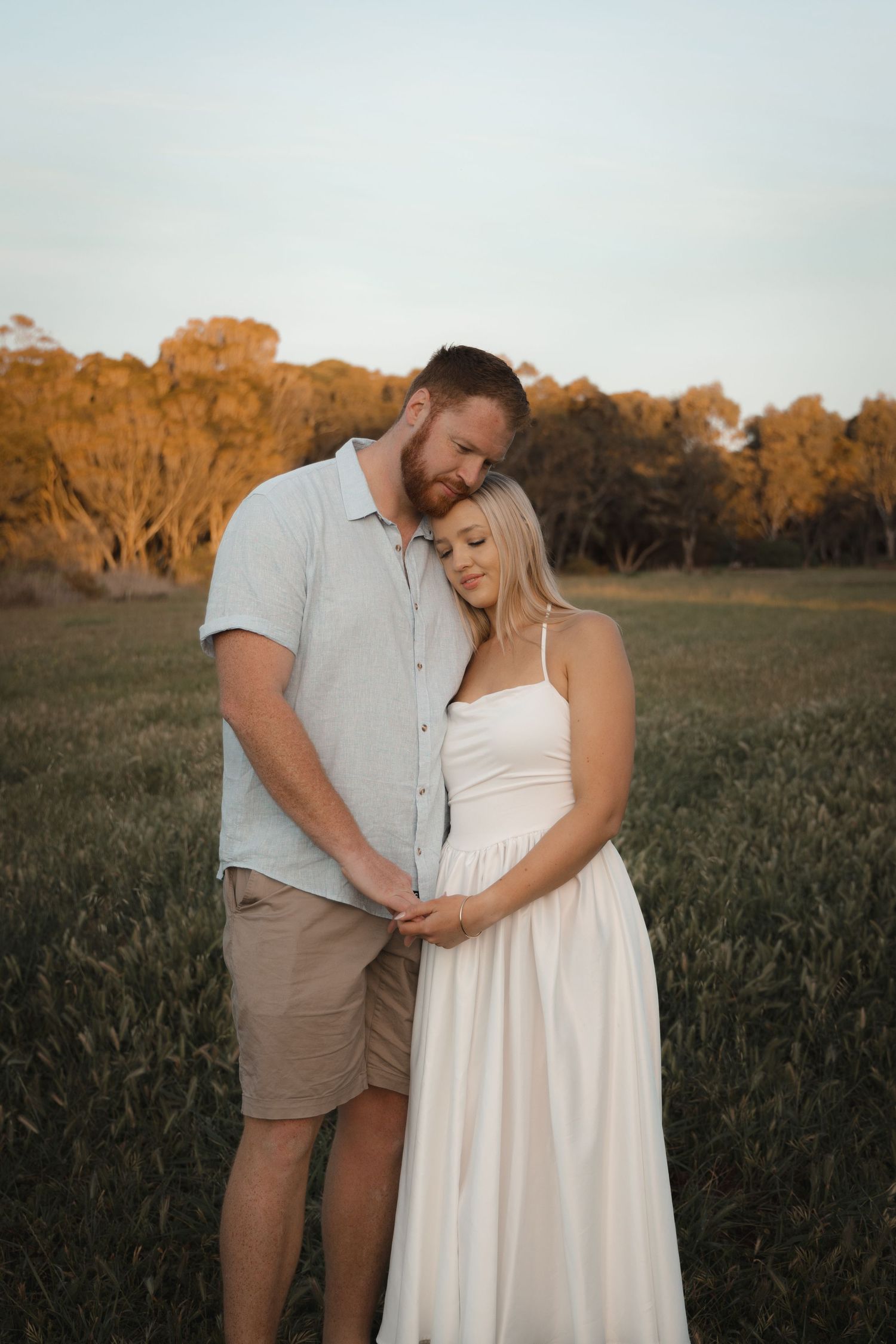 Couple sharing a tender maternity moment in a sunlit field, woman in white dress cradling baby bump at golden hour.