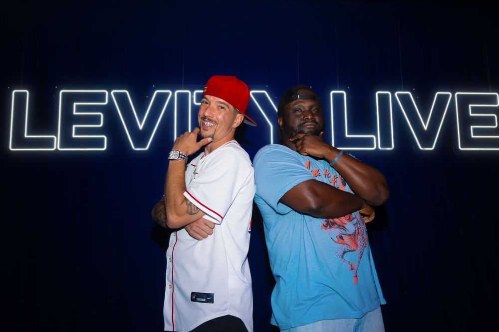 Two men pose back-to-back in front of a neon Levity Live sign, one wearing a red cap and baseball jersey.