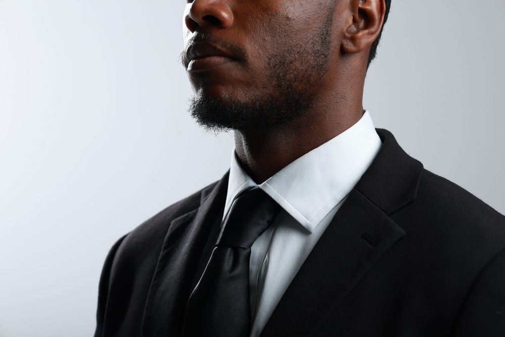 A professional Black man in a black suit and tie with a white dress shirt, photographed against a light gray background.
