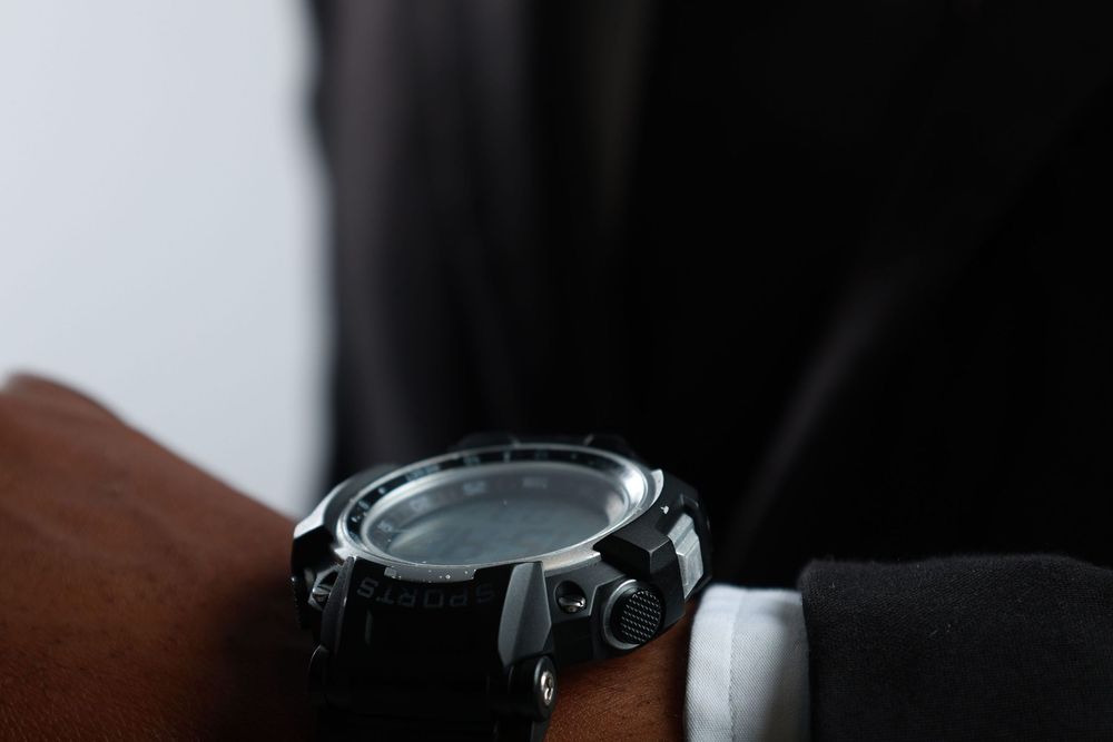 Close-up of a large black digital watch on a dark-skinned wrist, worn with a white shirt and black jacket.