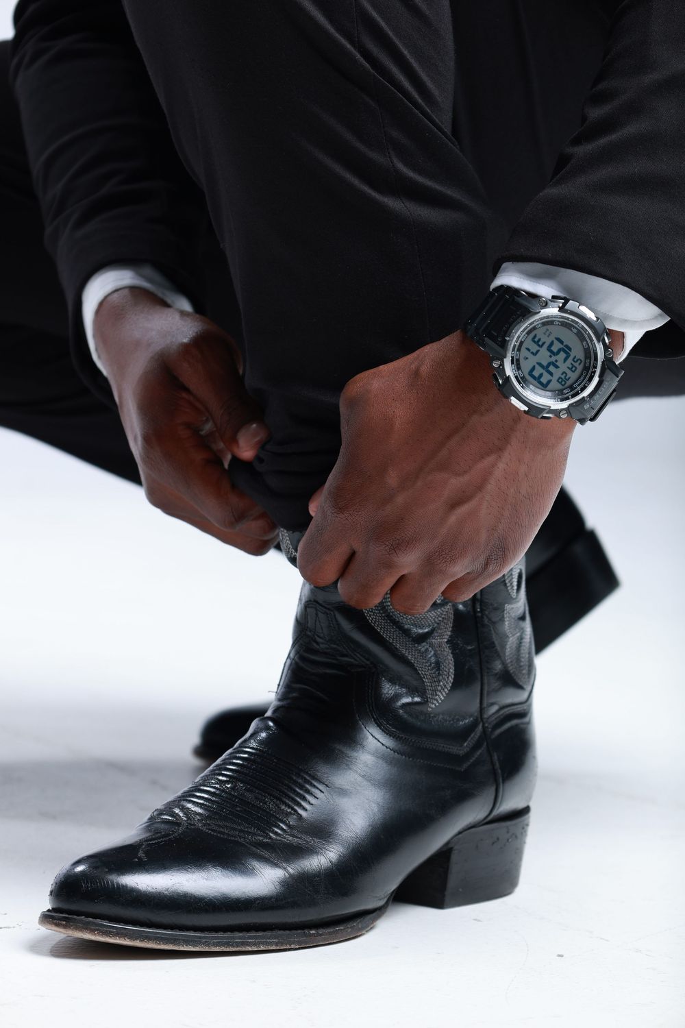 Man in black suit putting on black leather cowboy boots while wearing a digital watch.