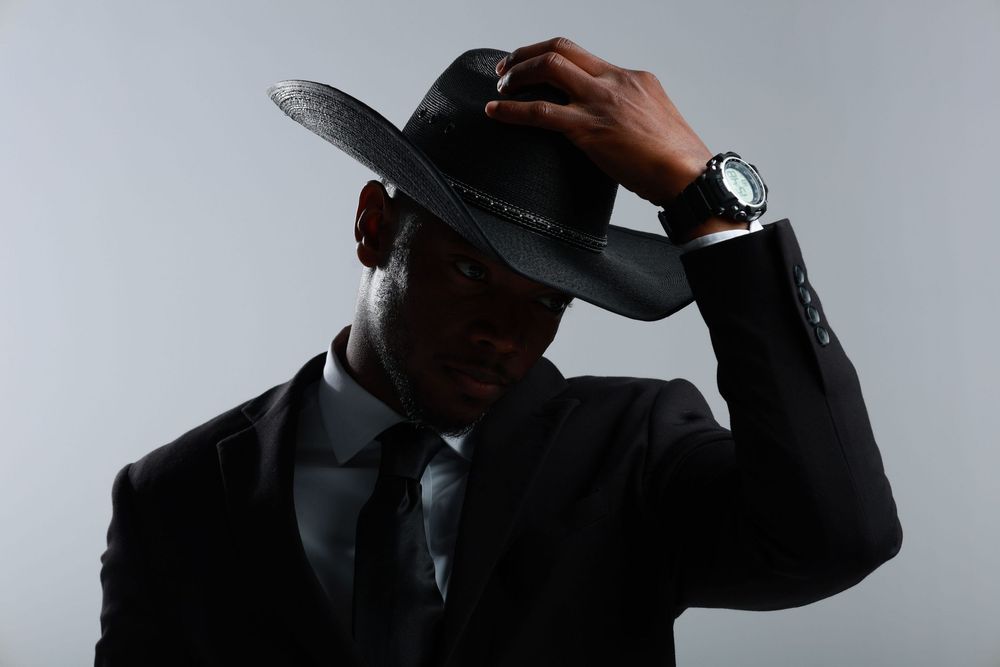 A stylish Black man in a black suit and tie adjusts his wide-brim fedora hat, wearing a silver watch.