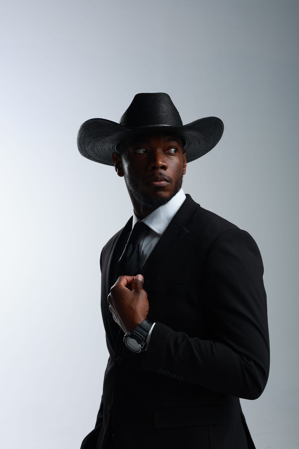 A stylish Black man in a black suit and cowboy hat poses confidently against a light gray background.