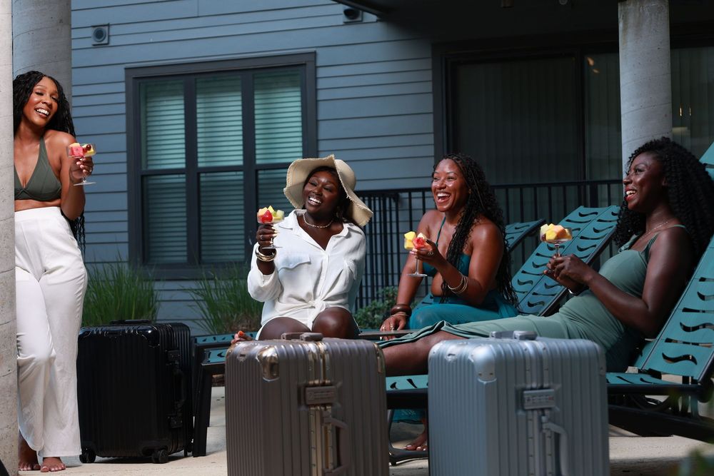 Four women laughing and enjoying drinks outdoors near luggage on a sunny day.