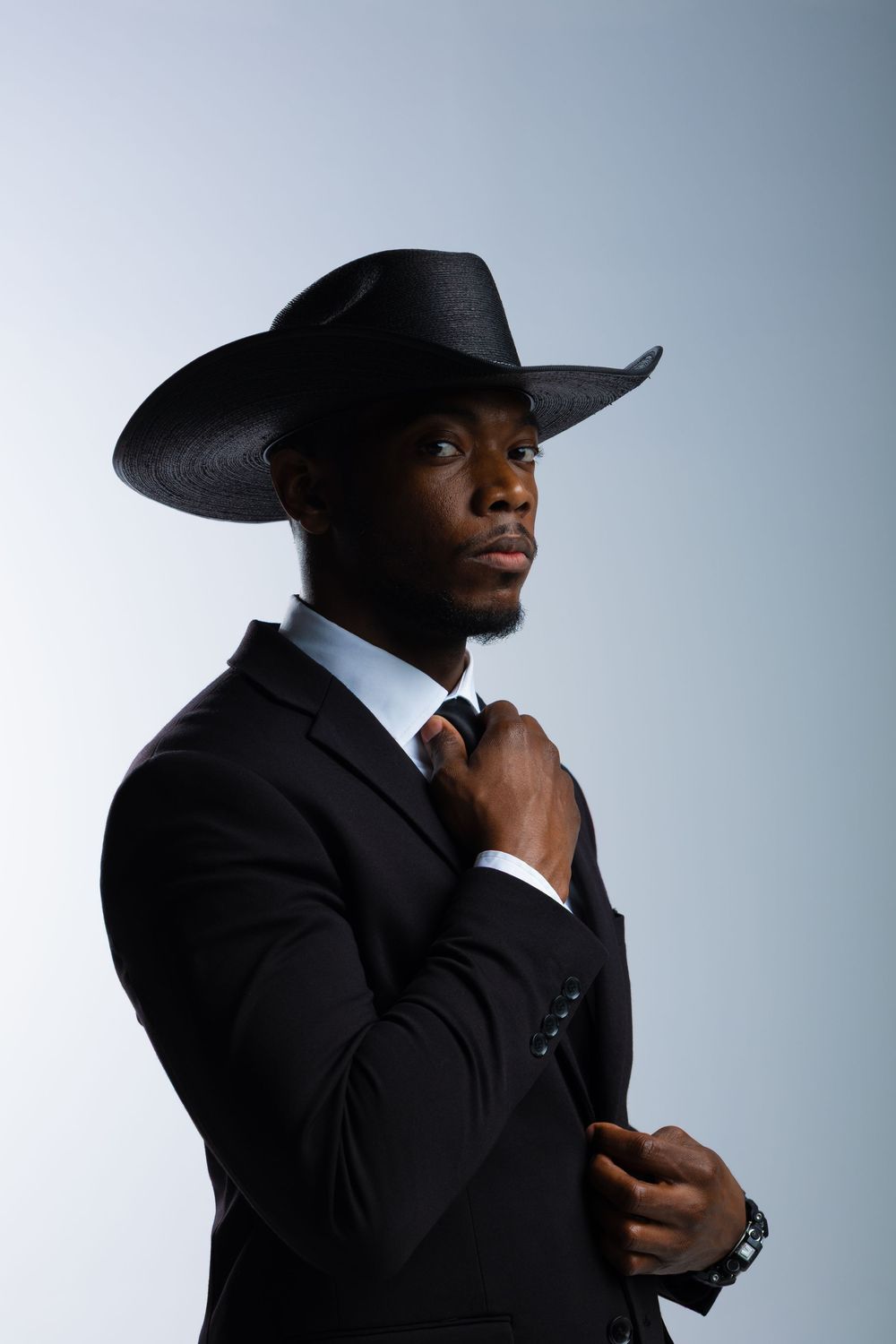 A stylish Black man in a black suit and cowboy hat adjusts his tie against a light gray background.