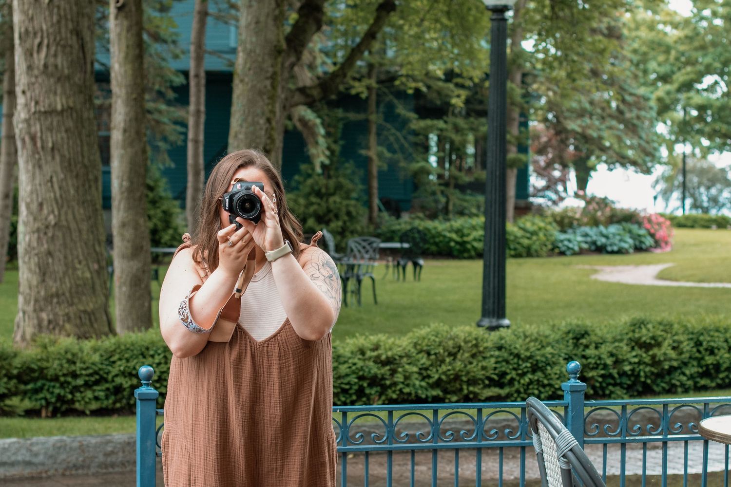 A photographer stands in front of a garden fence using a camera on a bright summer day.