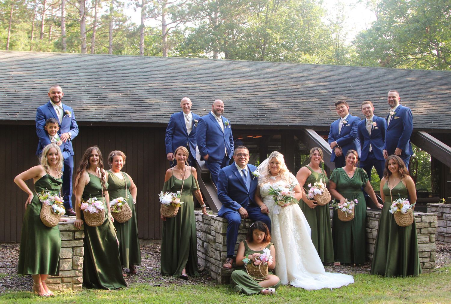 Wedding party in blue suits and green dresses poses for formal group photo outside rustic building.