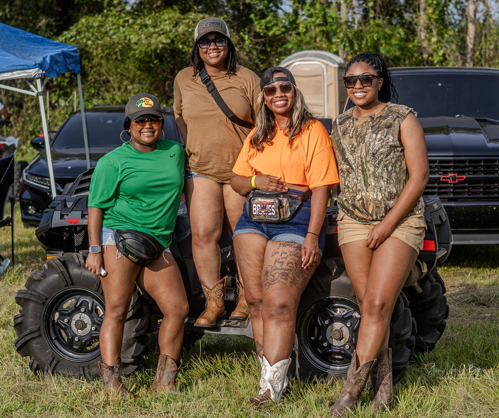 Four friends in casual outdoor wear pose together in front of an ATV on grassy terrain on a sunny day.