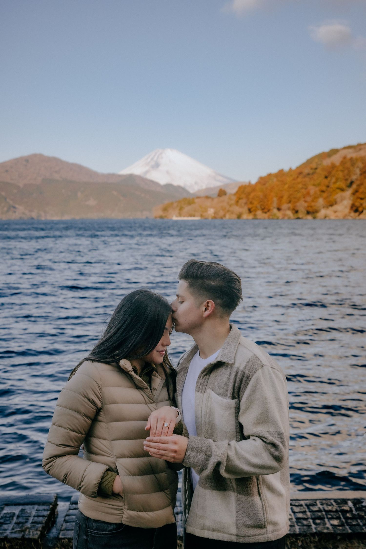 Two people in winter coats share a romantic moment by a lake with Mount Fuji and autumn foliage in the background.