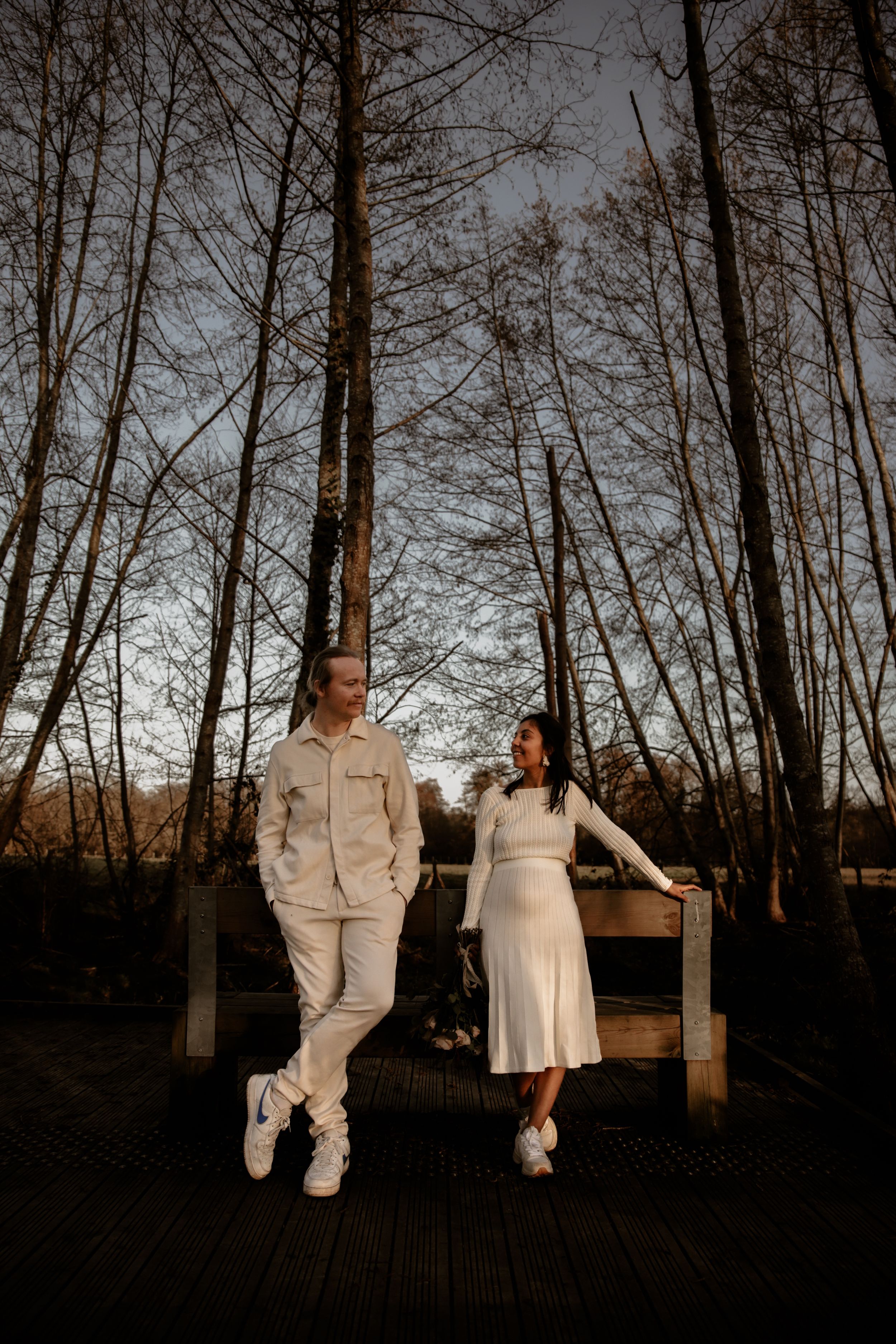 A couple in matching white outfits holds hands on a wooden boardwalk surrounded by tall trees at dusk.