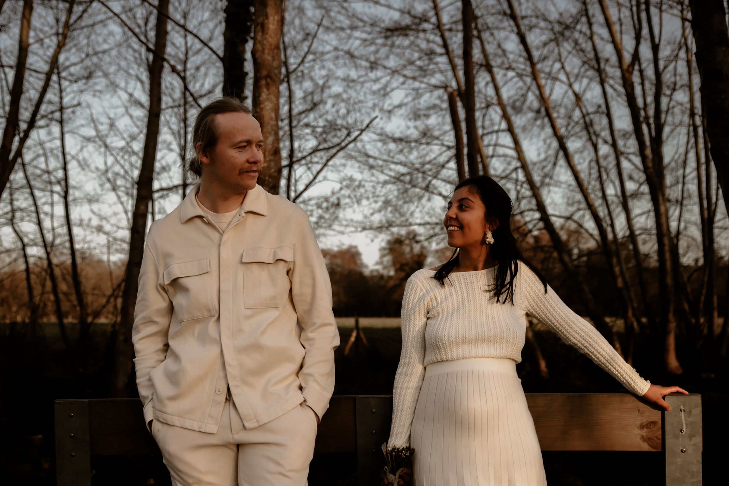 Couple in matching white outfits standing on wooden bridge in forest setting.