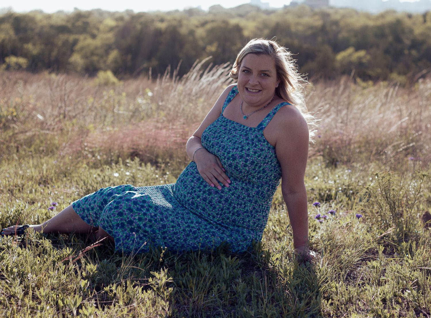 A person in a blue floral dress sits in a sunny meadow with tall grass and golden sunlight.