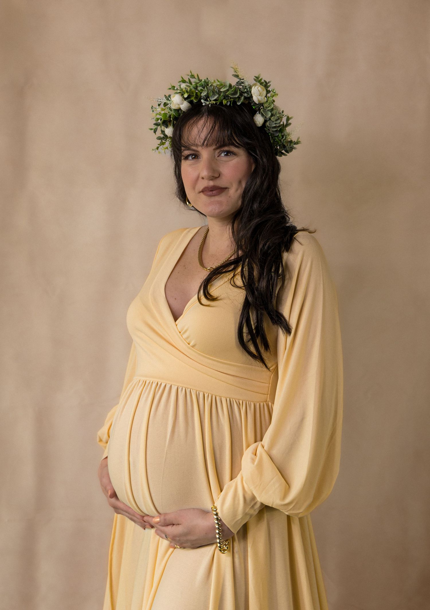 A person wearing a floral crown and yellow empire waist maternity dress poses against a beige backdrop.