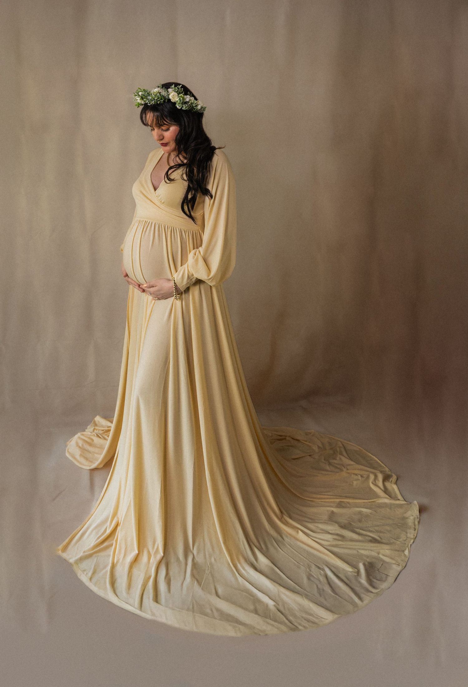 Studio maternity portrait with woman in cream gown and floral crown against neutral background.