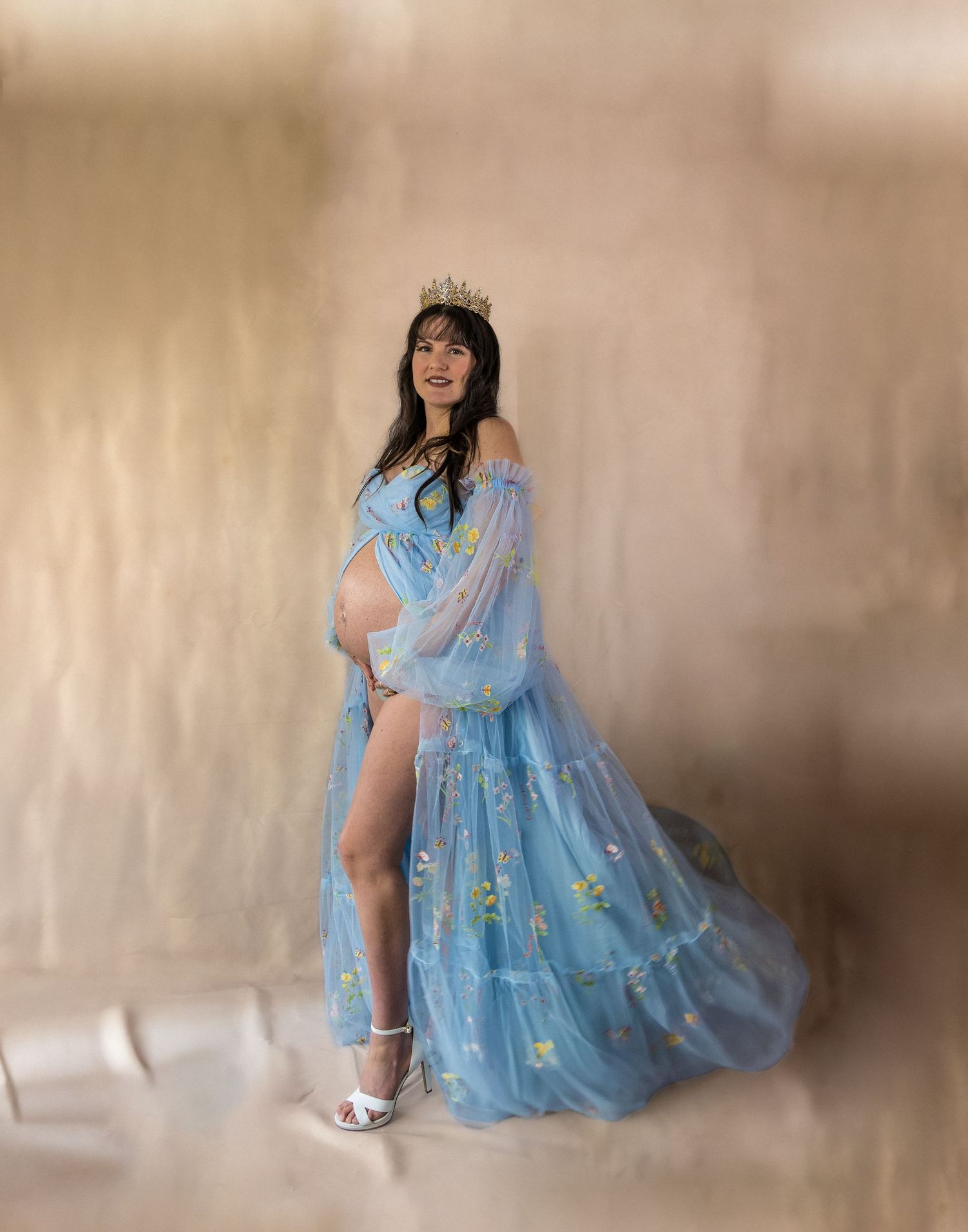 Ethereal maternity portrait in flowing blue gown with floral appliques against soft beige background.